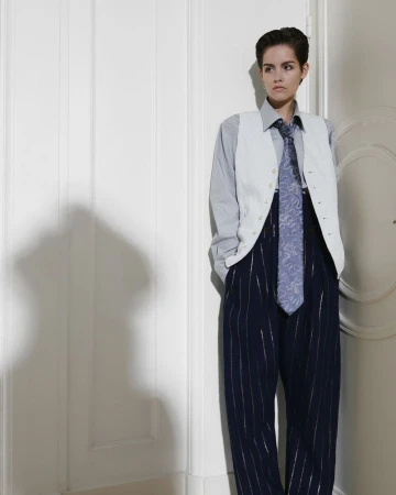 Woman with short hair wearing a vest, shirt, tie, and pinstripe pants stands indoors with a serious expression, shadowed by a white wall.