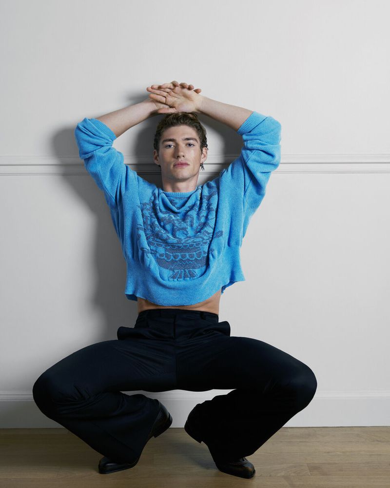 Male model with hands on head, wearing a turquoise sweater and black pants, sitting on the floor against an off-white wall in a photo studio.