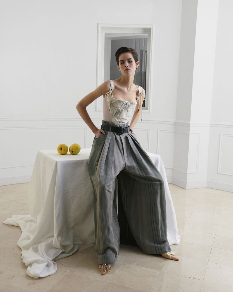 A woman poses indoors near a table wearing a corset top and wide-leg striped pants, bathed in soft lighting near a window.