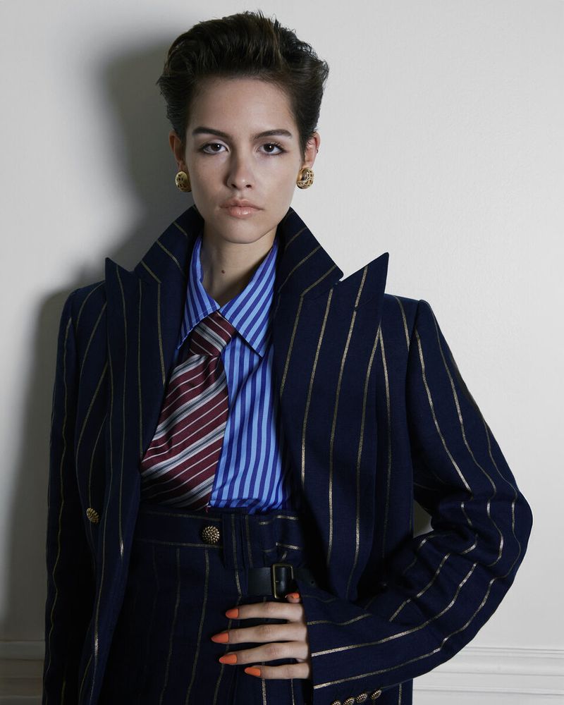 Adult female poses indoors wearing a striped blazer, blue shirt, and tie, accessorized with gold earrings. Portrait framing.