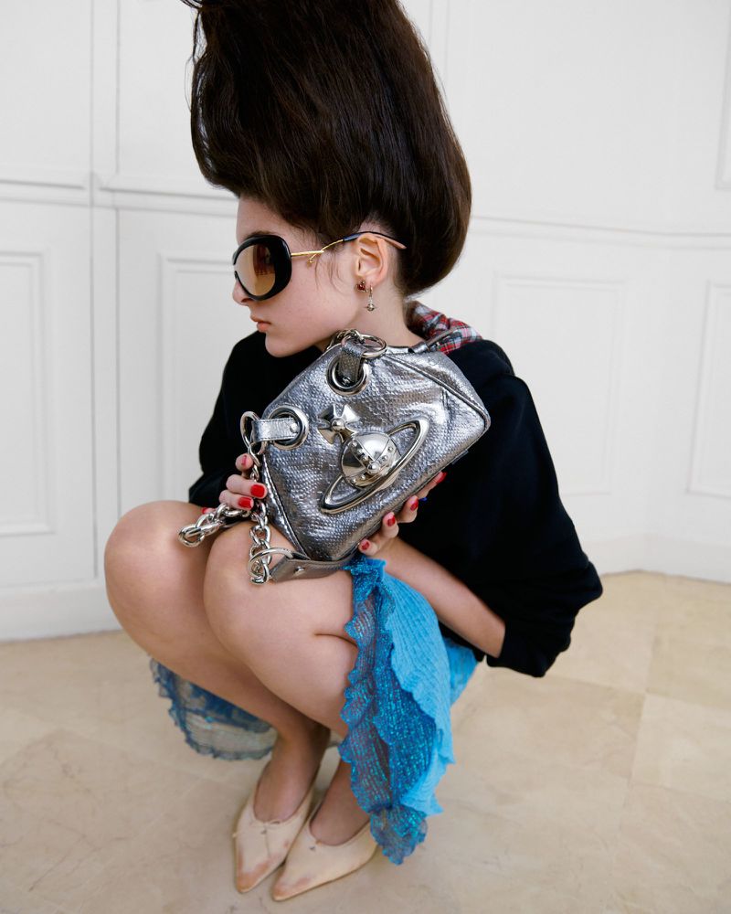 A young woman with high hair style and sunglasses, holding a silver Vivienne Westwood purse while squatting indoors.