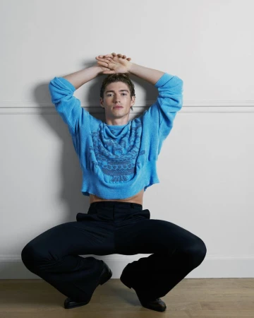 Model Valentin Humbroich crouching against a white wall in a cropped light blue knit sweater and tailored black trousers.