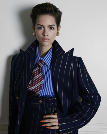 Model with short dark hair wearing an oversized navy pinstripe suit, blue striped shirt, and patterned tie against a pale backdrop.