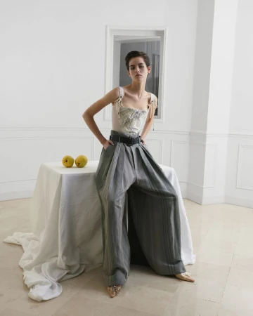 Model wearing a patterned corset top and oversized grey pleated trousers posing beside a white draped table with two yellow fruit.