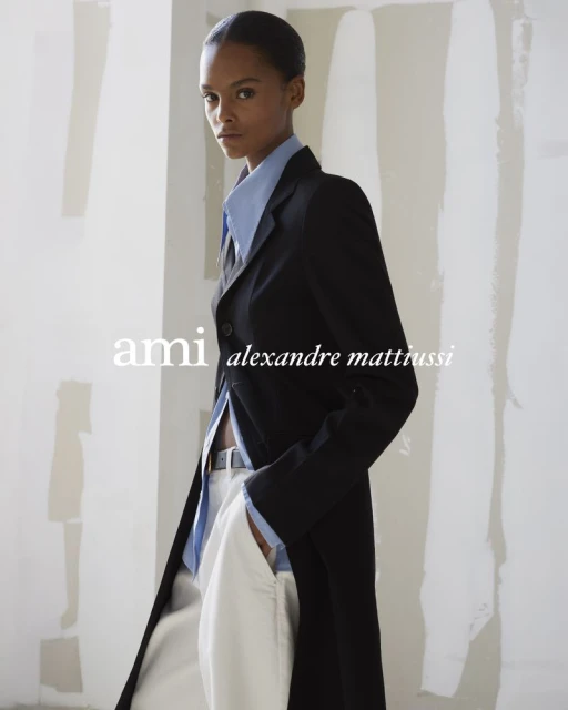 A woman in a light blue collared shirt and a black coat poses in front of a white background. Fashion editorial shot.