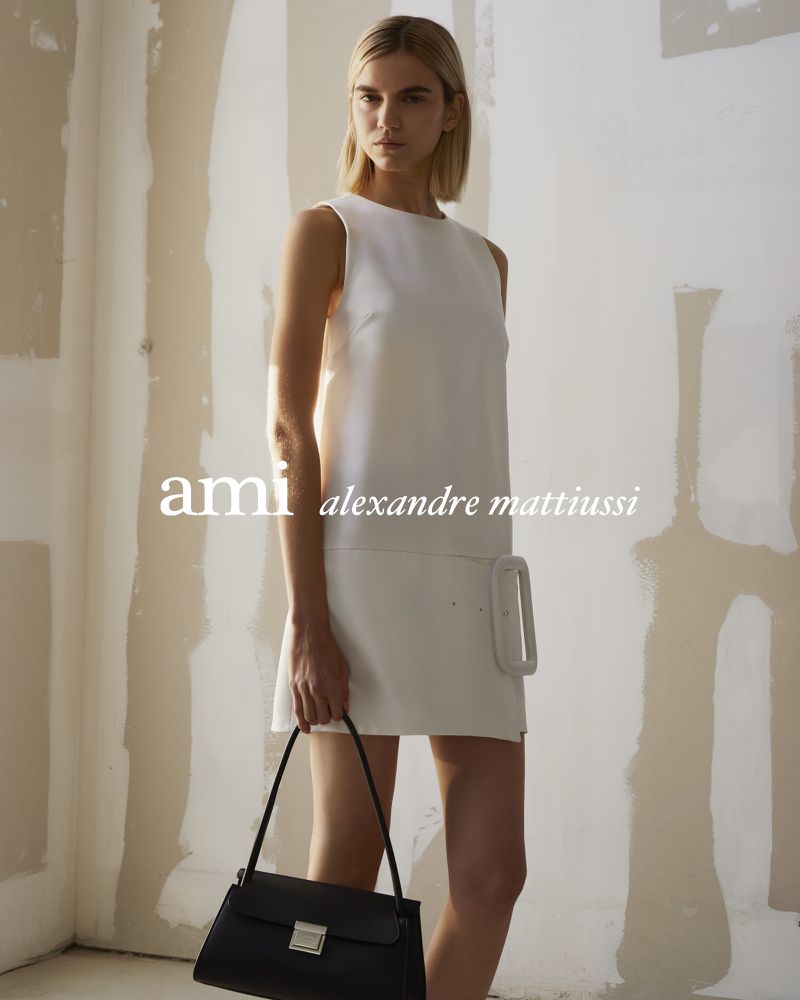 An adult female fashion model poses indoors wearing a sleeveless white mini dress and holding a black purse against a textured wall.