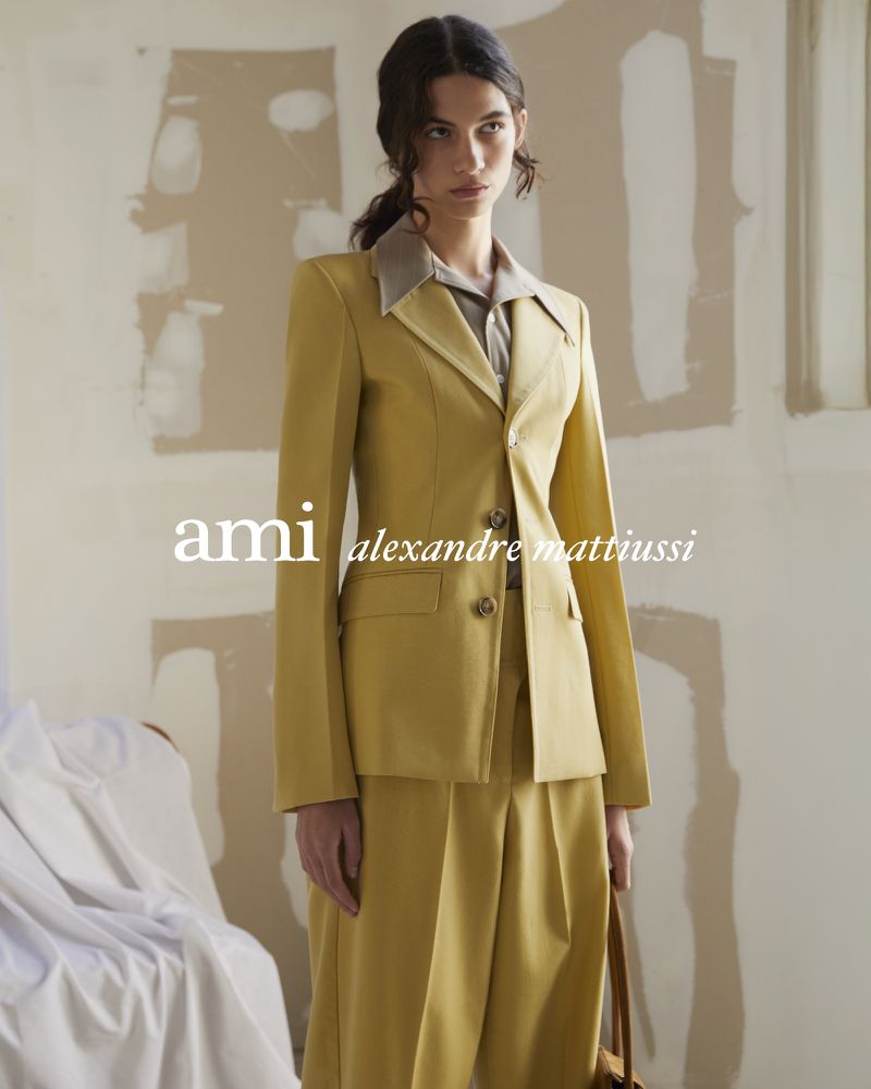 An adult female model poses indoors wearing a tailored yellow suit with wide-leg pants, alongside a tan handbag, against a neutral background.