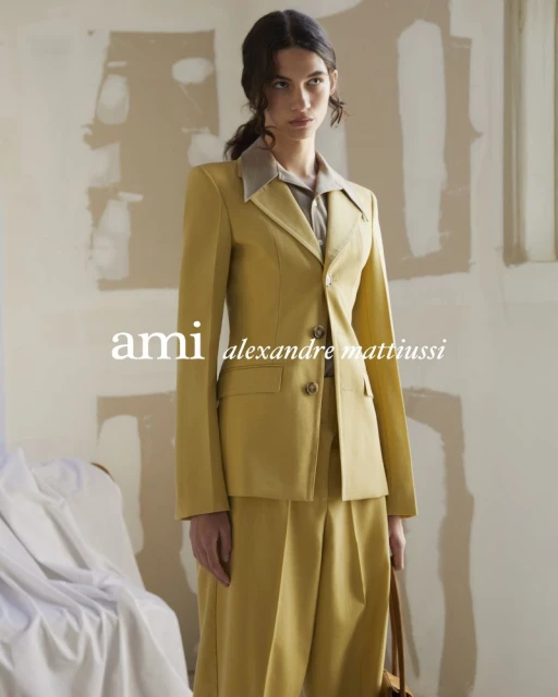 An adult female model poses indoors wearing a tailored yellow suit with wide-leg pants, alongside a tan handbag, against a neutral background.