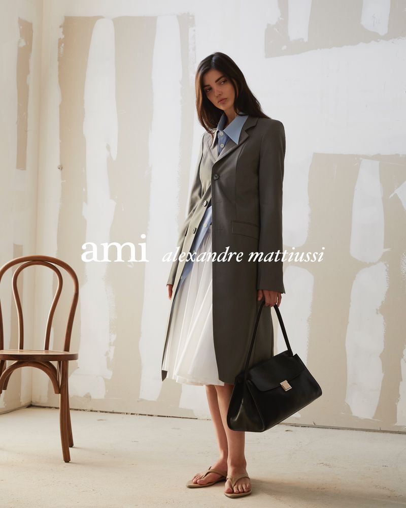 Model in grey coat and blue shirt holding a black handbag standing against a rustic wall.