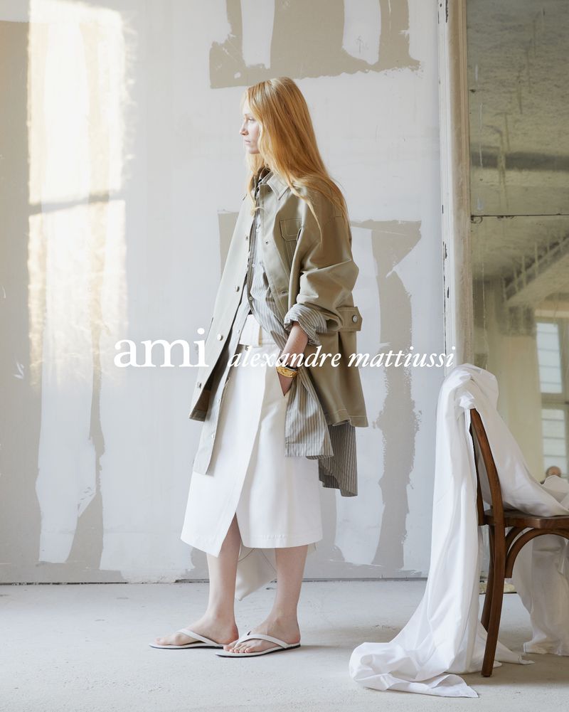 A woman with long red hair poses in a light-filled space wearing a trench coat, top, and skirt in a fashion editorial style.