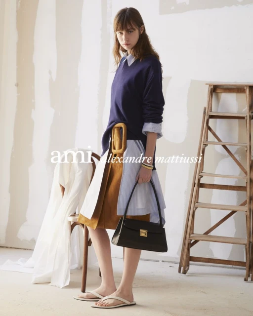 Fashion shot of a brunette woman in a layered outfit with a blue sweater, striped dress, brown belt, black purse, and beige sandals.