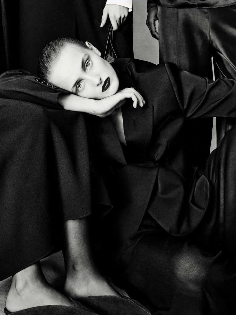 Black-and-white fashion shot of a reclining woman in a dark blazer, surrounded by three other models in black outfits against a neutral backdrop.