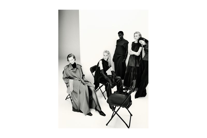 Black and white fashion shot of five female fashion models in dresses and a trench coat, seated and standing against a white backdrop.