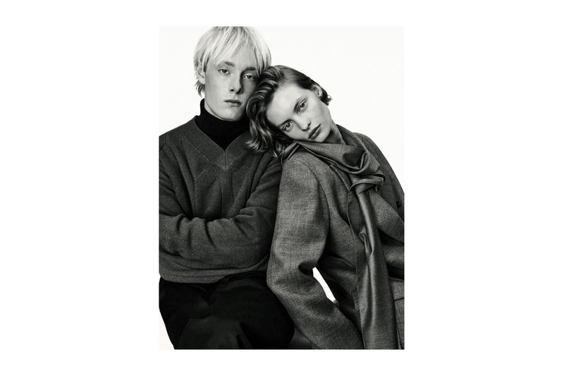 Black and white portrait of a young couple in gray sweaters and a gray jacket with a scarf against a white backdrop in a studio.