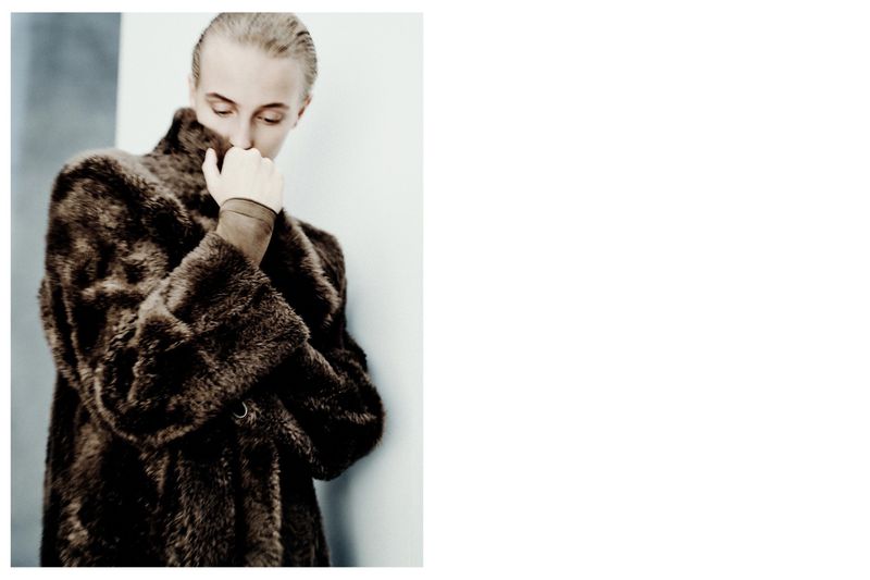 Adult woman in a brown fur coat leans against a wall with her head bowed and her hand covering her mouth in a studio setting.