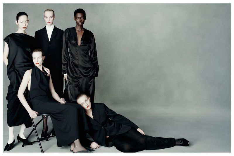 Fashion editorial image of five diverse women wearing black outfits in a studio setting, striking poses against a neutral gray backdrop.