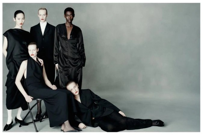 Fashion editorial image of five diverse women wearing black outfits in a studio setting, striking poses against a neutral gray backdrop.