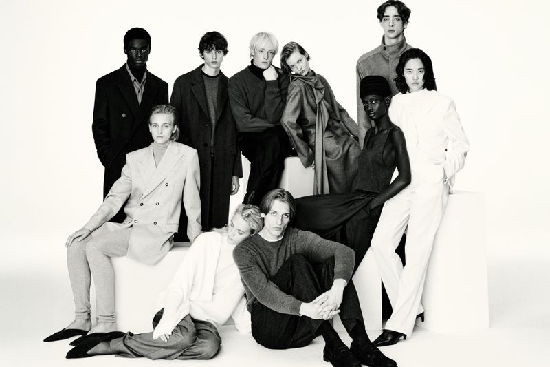Black and white studio shot of a group of nine diverse models in coats, sweaters, pants, and blazers posed on white blocks.