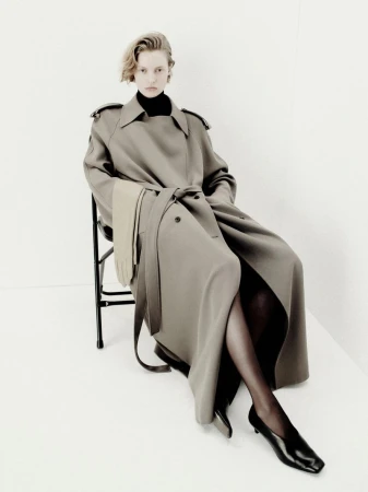 A woman with blond hair sits in a black chair wearing a gray trench coat, black turtleneck, tights, and black heeled shoes against white background.