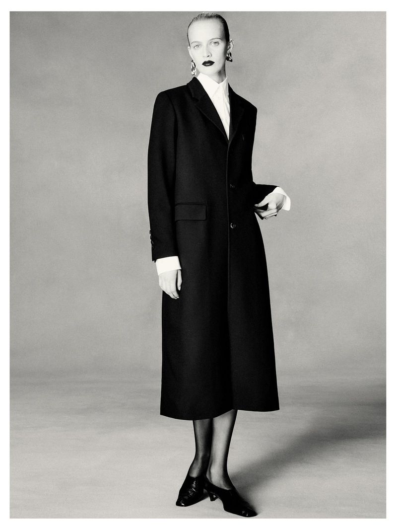 Elegant monochrome portrait of a fair-skinned woman in a black coat, white blouse, stockings, and heels against a grey backdrop.