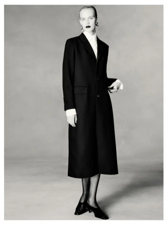 Elegant monochrome portrait of a fair-skinned woman in a black coat, white blouse, stockings, and heels against a grey backdrop.