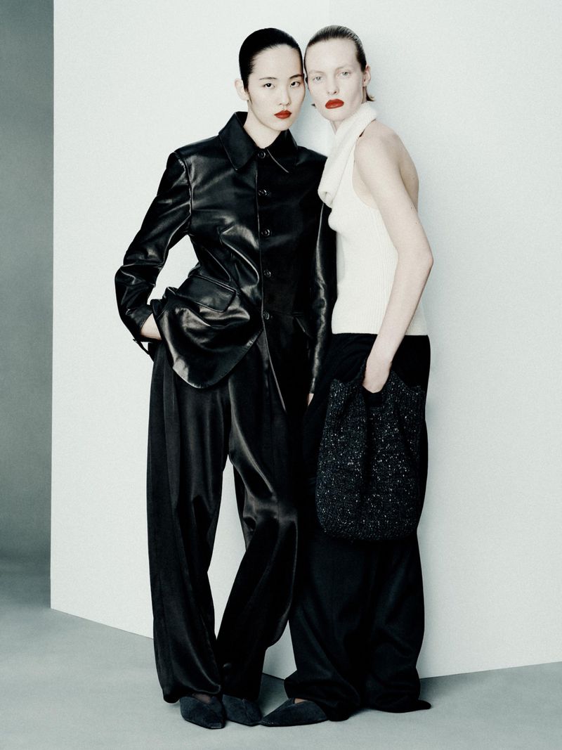 Two adult women pose in a photo studio, wearing dark and light colour outfits, featuring a leather jacket, wide-leg pants, and a small bag.