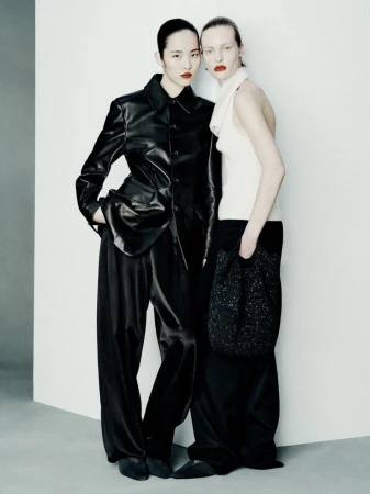 Two adult women pose in a photo studio, wearing dark and light colour outfits, featuring a leather jacket, wide-leg pants, and a small bag.
