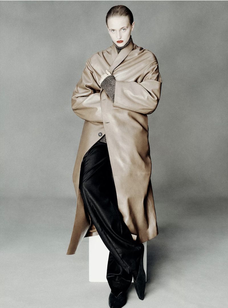 Model wearing an oversized camel leather coat, grey knit turtleneck, and black trousers posing against a grey studio backdrop.
