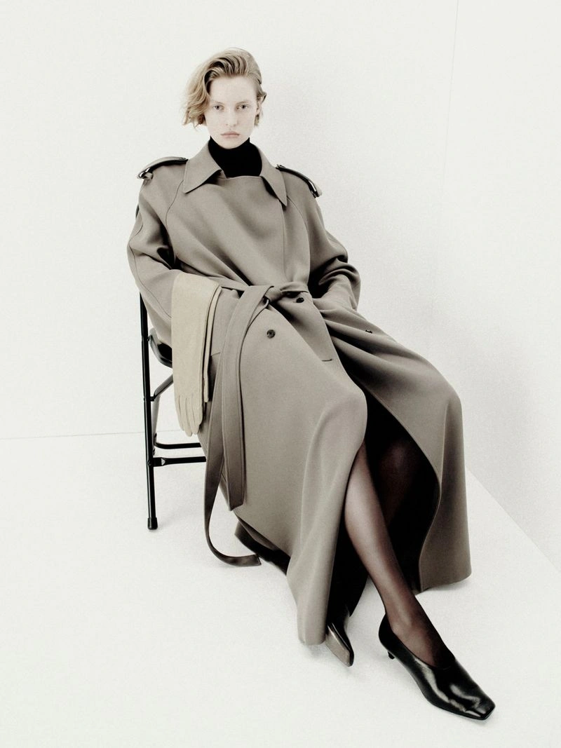 Model seated on a black chair wearing an oversized taupe belted coat, black heels, and beige gloves against a white studio corner.