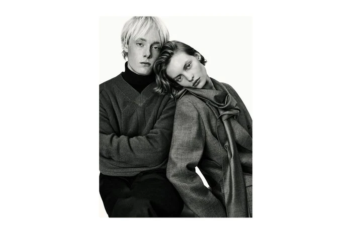 Monochrome studio portrait of a man in a knit sweater and a woman in a tailored wool jacket and scarf leaning on his shoulder.