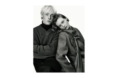 Monochrome studio portrait of a man in a knit sweater and a woman in a tailored wool jacket and scarf leaning on his shoulder.