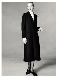 Model wearing a long black coat over a white shirt against a neutral studio backdrop