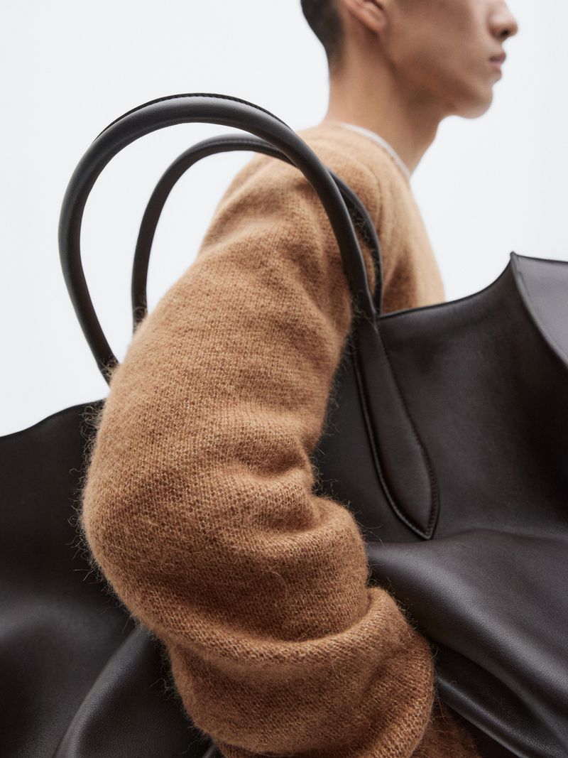 Man wearing a tan sweater carrying a dark brown leather tote bag in a minimalist fashion shot with a bright background.