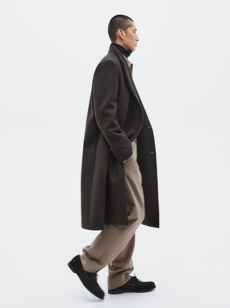 Stylish Asian male model in brown coat, tan pants, black shoes, walking in a minimalist white photo studio with soft, neutral lighting.
