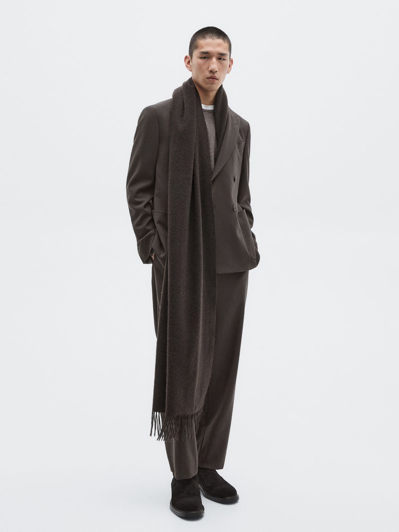 Studio shot of a young adult male fashion model in a brown suit and scarf against a white background; full-length view with neutral expression.