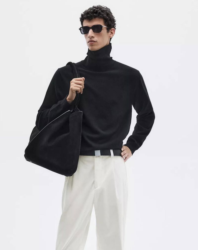 Male model wearing a black turtleneck sweater, white trousers, and dark sunglasses carrying a black shoulder tote against a white backdrop.