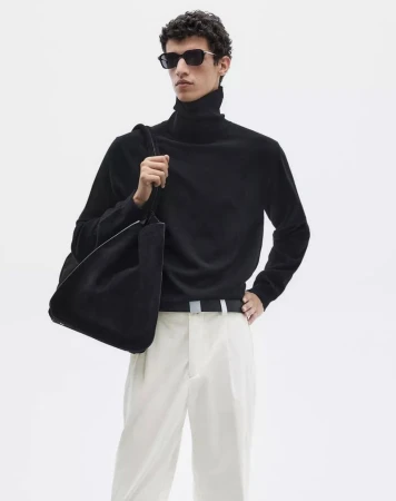 Male model wearing a black turtleneck sweater, white trousers, and dark sunglasses carrying a black shoulder tote against a white backdrop.