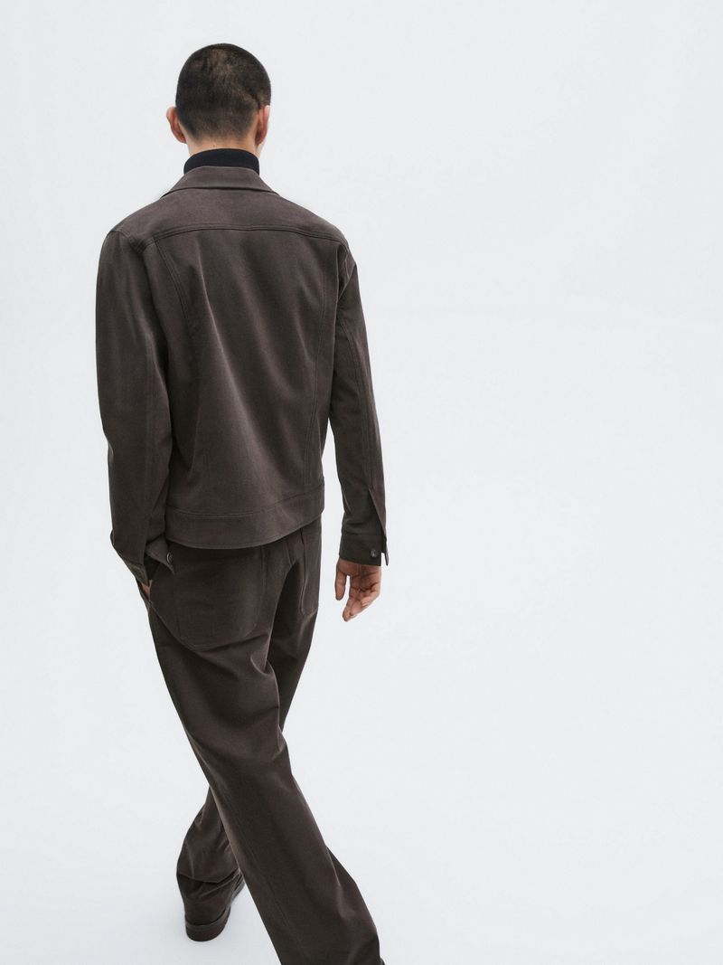 Rear view of a man walking away in a dark brown jacket and matching trousers against a white seamless background.