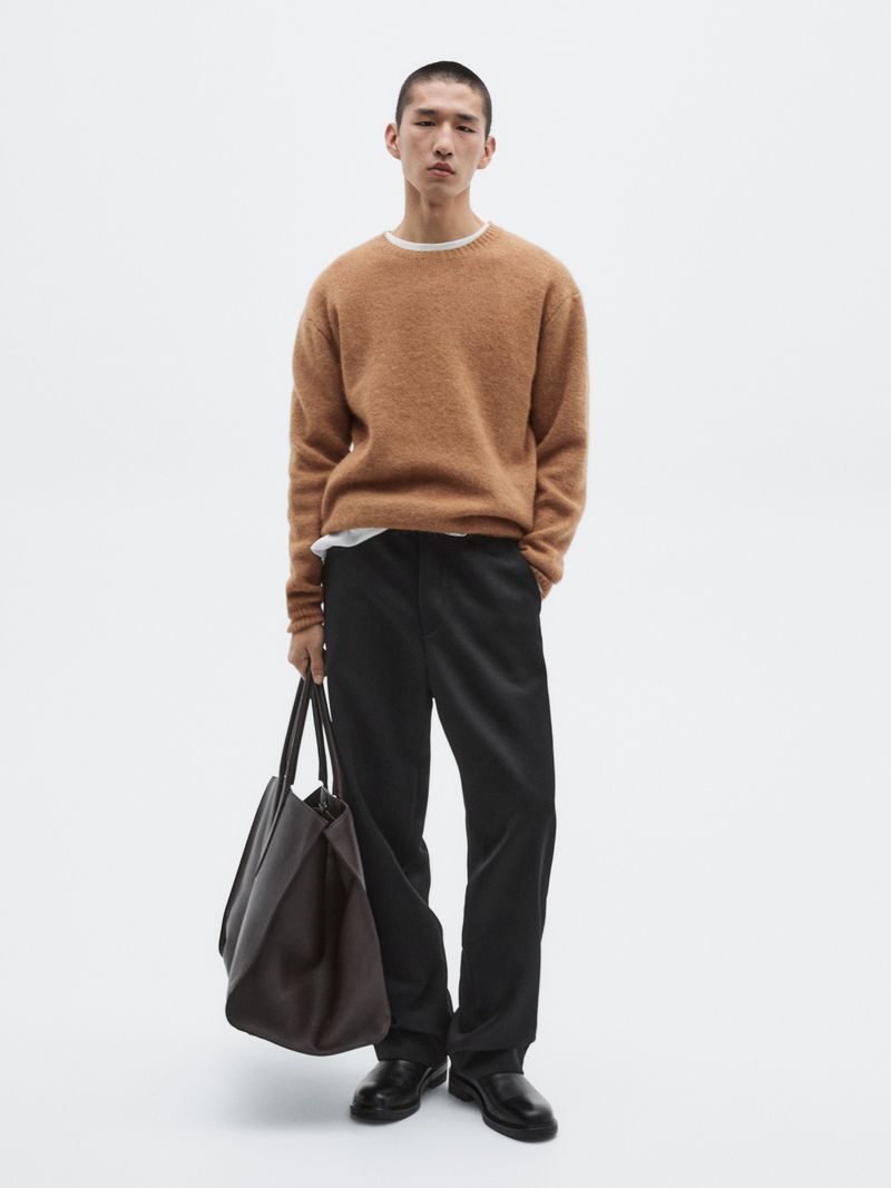 Model Long Li posing in a brown textured knit sweater, black trousers, and leather shoes while holding a dark tote bag against a white backdrop.