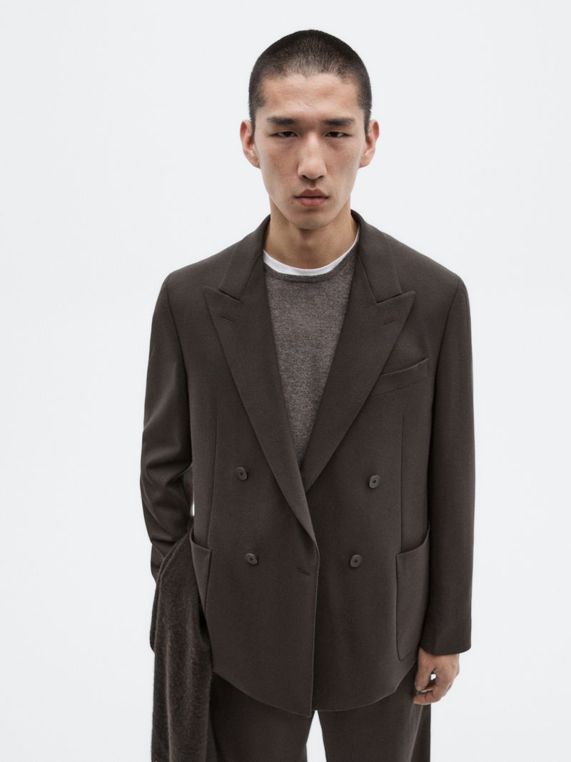 A stylish Asian man is showcased in a studio shot, sporting a double-breasted brown suit and a grey sweater against a clean white backdrop.