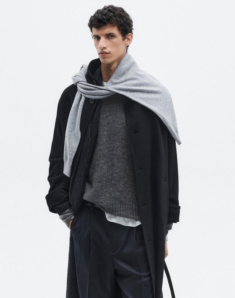 Male fashion model in a black coat and gray scarf poses in a studio against a white backdrop, showcasing neutral, elegant menswear.
