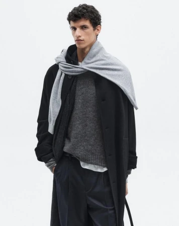 Male fashion model in a black coat and gray scarf poses in a studio against a white backdrop, showcasing neutral, elegant menswear.