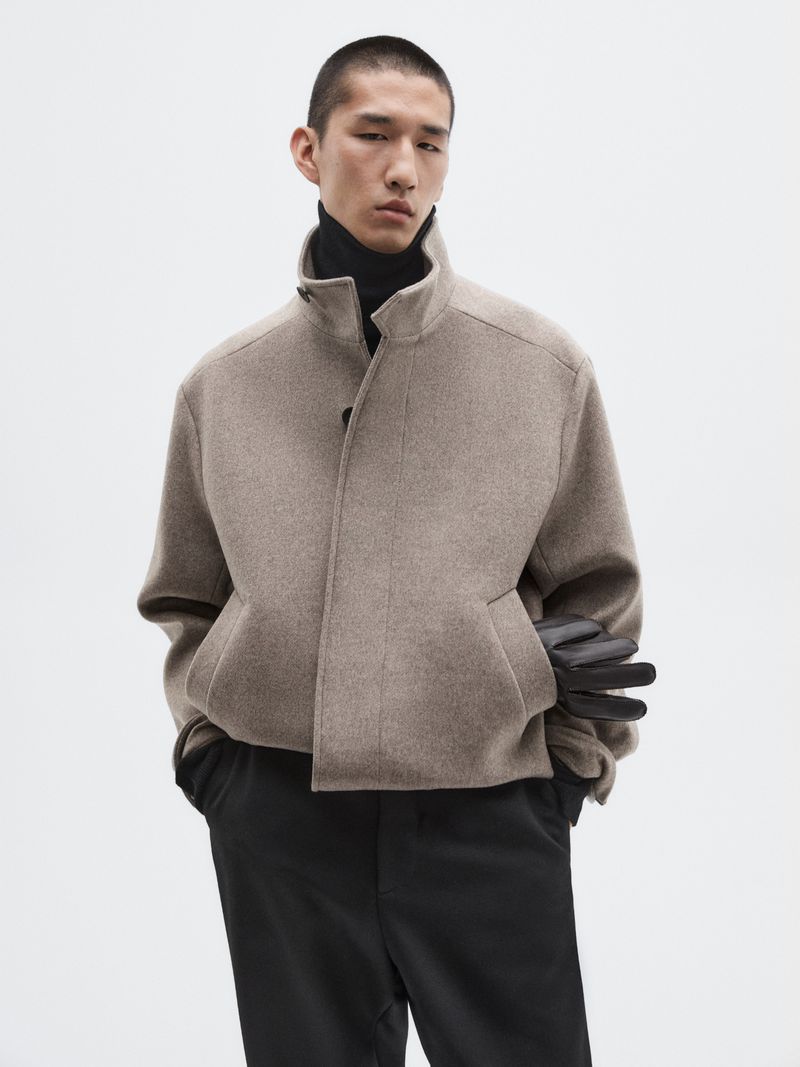 Man poses indoors wearing a wool jacket and black pants in a fashion shot. The background is white, and the lighting is neutral.