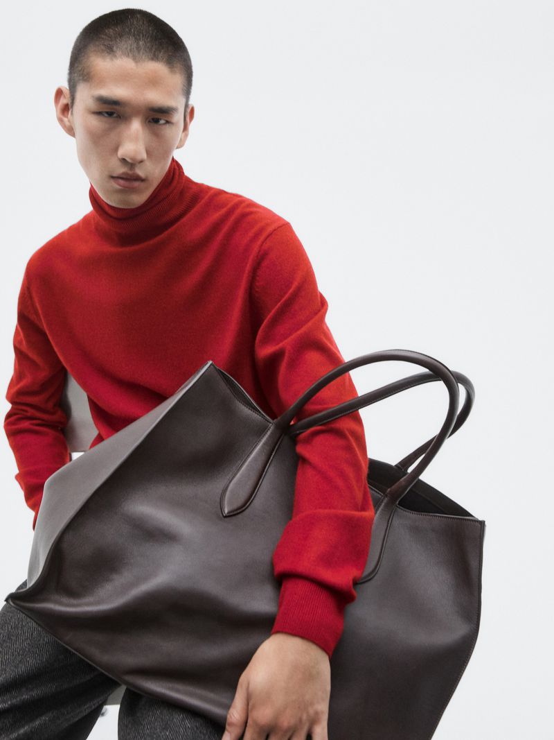 Fashion shot of a model in a red turtleneck sweater holding a large brown leather tote bag against a bright white background.