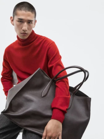 Fashion shot of a model in a red turtleneck sweater holding a large brown leather tote bag against a bright white background.