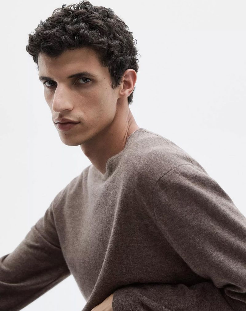 Close-up shot of a young adult male model with curly dark hair wearing a long-sleeve brown cashmere sweater against a white background.