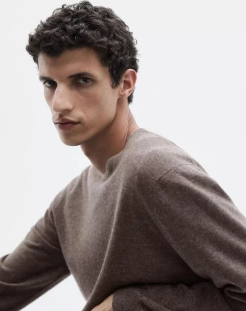 Close-up shot of a young adult male model with curly dark hair wearing a long-sleeve brown cashmere sweater against a white background.