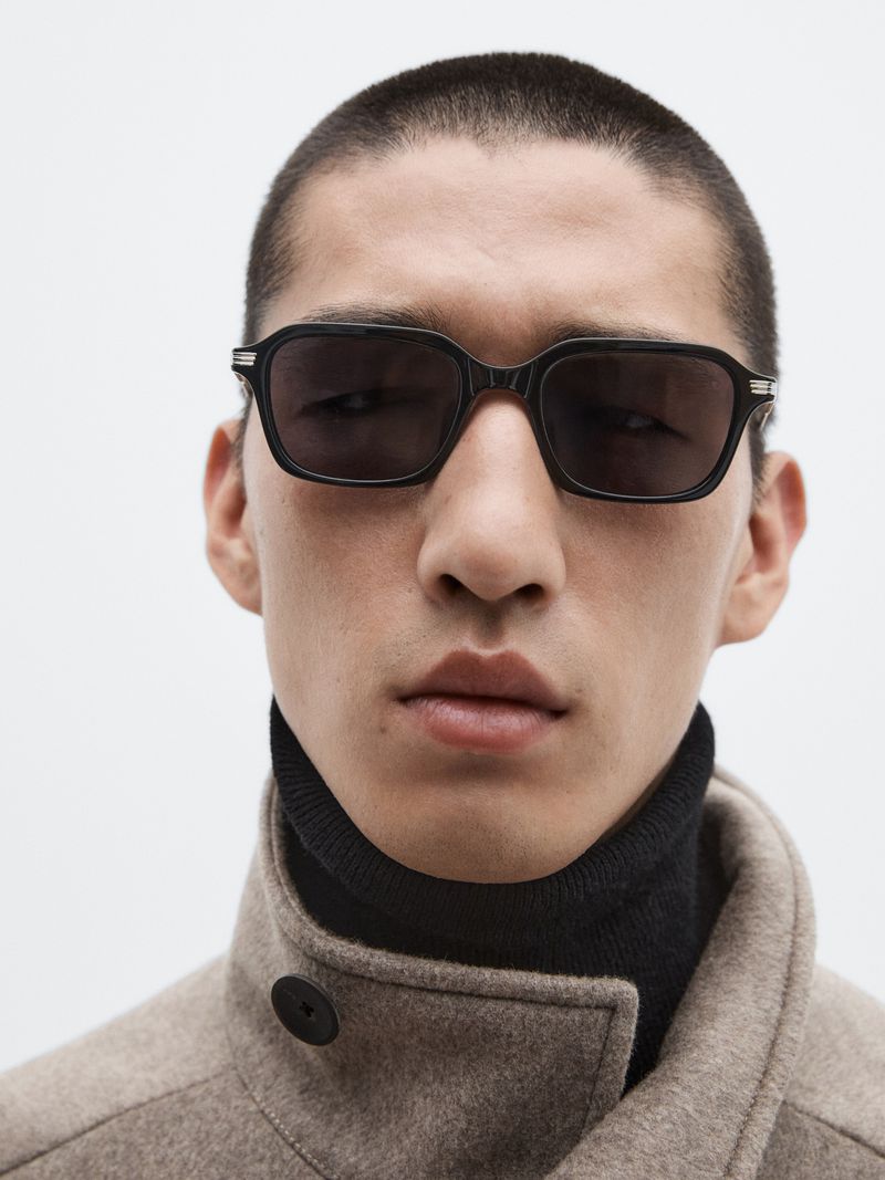 Close-up of a stylish adult male wearing sunglasses and a turtle neck, complemented by a wool coat against a plain white backdrop.