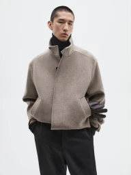 Model Long Li wearing a taupe wool zip jacket, black turtleneck, and black leather gloves against a white seamless backdrop.