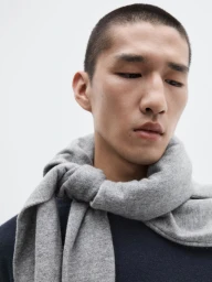 Model Long Li wearing a grey knit sweater tied around his neck over a dark blue top against a white studio background.